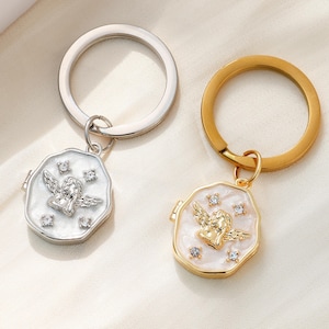 Könnte beinhalten: Zwei Schlüsselanhänger mit einem Engelscharme, einer mit einem silbernen Schlüsselring und einem weißen Emaille-Anhänger, der andere mit einem goldenen Schlüsselring und einem weißen Emaille-Anhänger. Beide Anhänger zeigen einen kleinen Engel mit Flügeln und Strasssteinen.