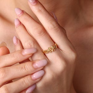 Könnte beinhalten: Ein goldfarbener Ring mit einem zentralen Buchstaben M als Blickfang, flankiert von einem Band im Kettenstil. Das Metall hat eine glatte, polierte Oberfläche und das Design enthält ein sich wiederholendes ovales Gliedermuster.