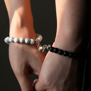 May include: Two bracelets on wrists, one white with a heart charm, the other black.