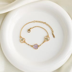 Op de afbeelding: Een gouden kettingarmband met drie hartvormige bedels. Elke bedel heeft een ander gedroogd bloemenmotief in tinten van paars, wit en groen.