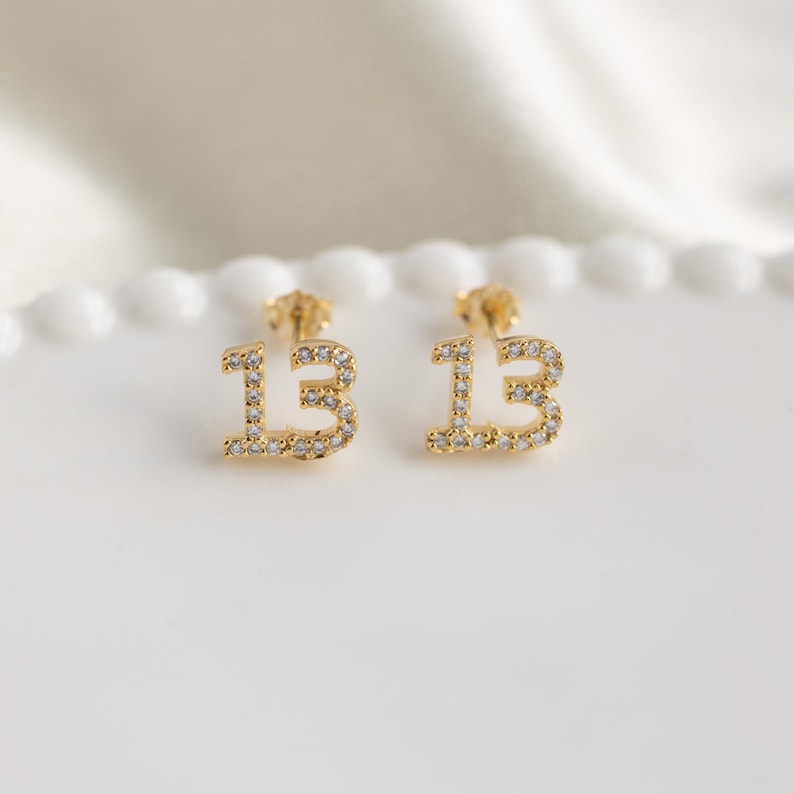 May include: A pair of gold earrings with the number 13 set with clear cubic zirconia stones.