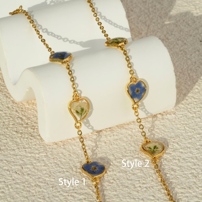 Op de afbeelding: Twee gouden kettingen met hartvormige hangers met geperste bloemen. De ketting aan de linkerkant heeft een witte bloem en de ketting aan de rechterkant heeft een blauwe bloem. De kettingen zijn ge&euml;tiketteerd met "Style 1" en "Style 2".