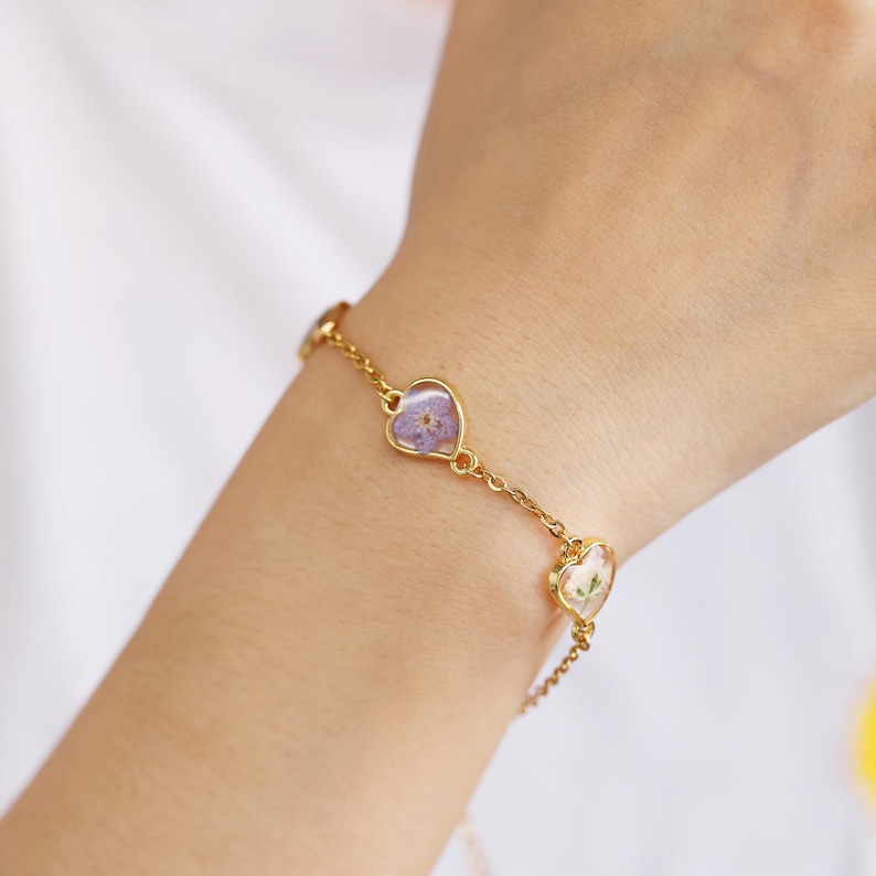 Op de afbeelding: Een gouden kettingarmband met drie hartvormige bedels. Elke bedel heeft een geperste bloem erin. De bloemen zijn paars, wit en groen.