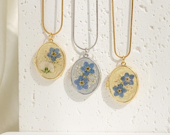 Könnte beinhalten: Drei goldene und silberne ovale Medaillon-Halsketten mit zarten Ketten. Jedes Medaillon zeigt ein gepresstes Blumenmotiv mit blauen und weißen Blüten.