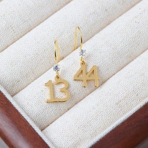 May include: A pair of gold-toned drop earrings featuring the numbers 13 and 44. Each earring is suspended from a curved hook and includes a small, clear, round faceted stone set directly above the numeral charm, creating a dangling design.
