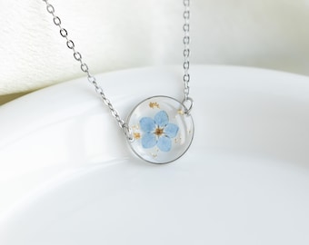 May include: A silver chain necklace with a round pendant featuring a delicate blue forget-me-not flower pressed inside clear resin.
