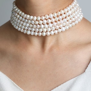 May include: A white pearl choker necklace with multiple strands of pearls. The necklace is made of small, round pearls and is worn by a person.