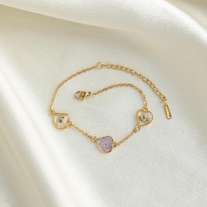 Op de afbeelding: Een gouden kettingarmband met drie hartvormige bedels. Elke bedel heeft een andere geperste bloem erin.