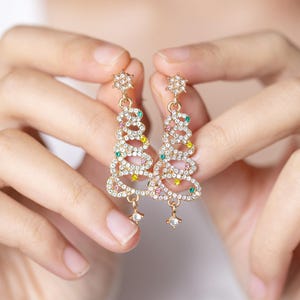 Puede incluir: Un par de pendientes colgantes de oro con forma de árbol de Navidad. Los pendientes están decorados con diamantes de imitación transparentes y de colores.