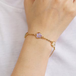 Op de afbeelding: Een gouden kettingarmband met drie hartvormige bedels. Elke bedel heeft een geperste bloem erin. Twee bedels hebben paarse bloemen en &eacute;&eacute;n bedel heeft een witte bloem.