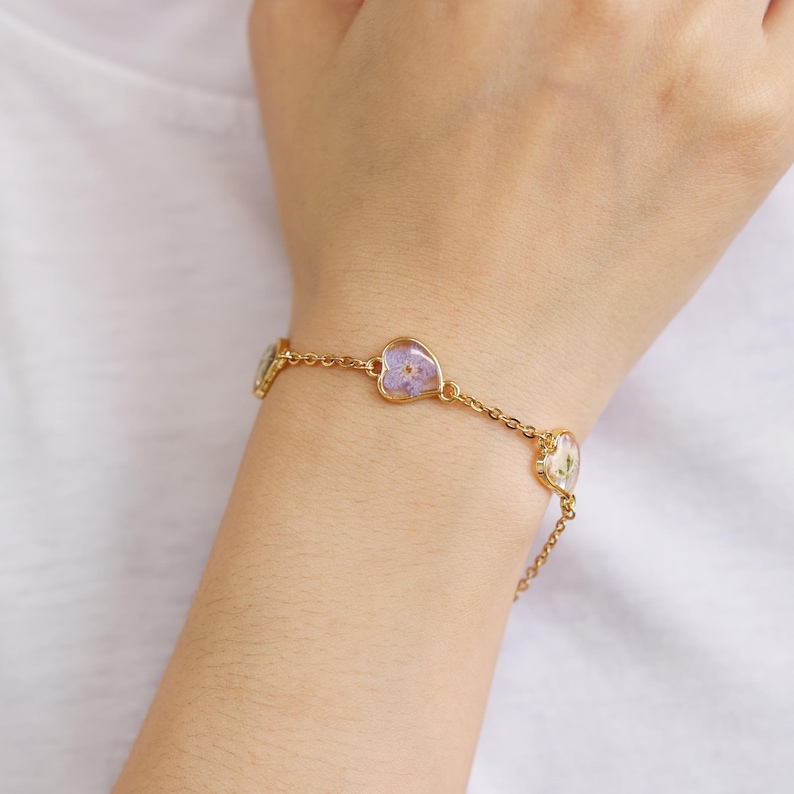 Op de afbeelding: Een gouden kettingarmband met drie hartvormige bedels. Elke bedel heeft een geperste bloem erin. Twee bedels hebben paarse bloemen en &eacute;&eacute;n bedel heeft een witte bloem.