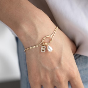 May include: A gold chain bracelet with a pearl charm and a letter 'B' charm. The bracelet is adjustable and has a knot design.