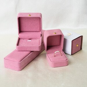 May include: Three pink velvet jewelry boxes with lids. The largest box is open and has a ring inside. The smallest box is closed and has a white label with the text "The Crown Jewels".