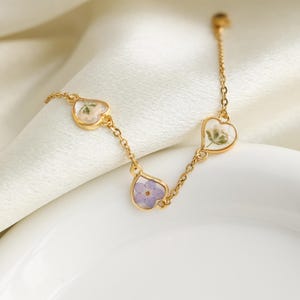 Op de afbeelding: Een gouden kettingarmband met drie hartvormige bedels. Elke bedel heeft een andere geperste bloem erin. De bloemen zijn wit, paars en een mix van wit en groen.