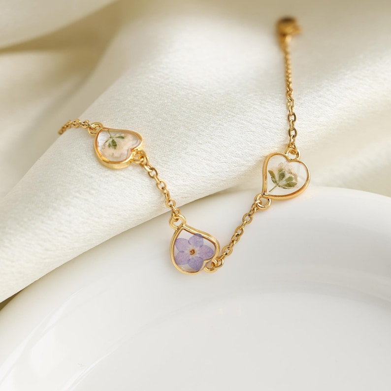 Op de afbeelding: Een gouden kettingarmband met drie hartvormige bedels. Elke bedel heeft een andere geperste bloem erin. De bloemen zijn wit, paars en een mix van wit en groen.