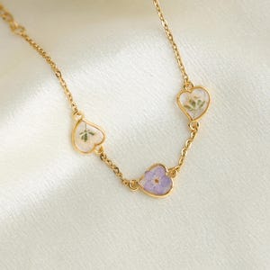 Op de afbeelding: Een gouden ketting met drie hartvormige hangers. Elke hanger heeft een ander gedroogd bloemenmotief, waaronder witte bloemen, paarse bloemen en groene bladeren.