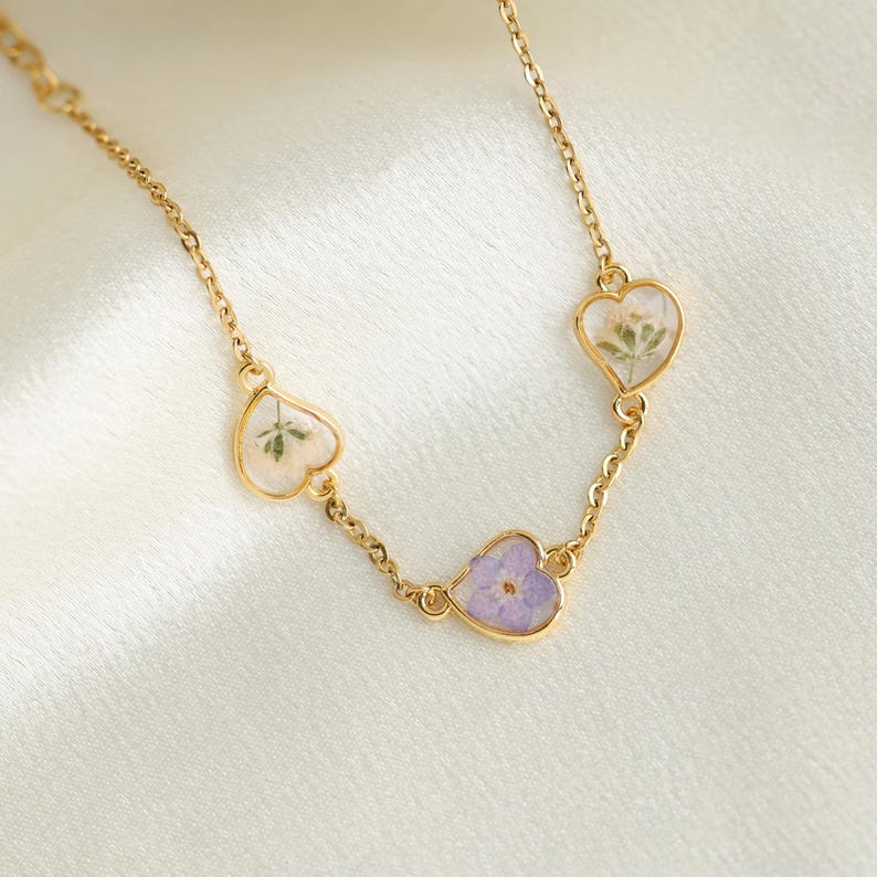 Op de afbeelding: Een gouden ketting met drie hartvormige hangers. Elke hanger heeft een ander gedroogd bloemenmotief, waaronder witte bloemen, paarse bloemen en groene bladeren.