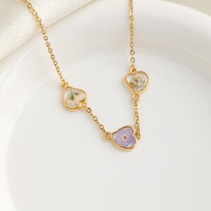 Op de afbeelding: Een gouden ketting met drie hartvormige hangers. Elke hanger heeft een andere geperste bloem erin. De bloemen zijn wit, groen en paars.