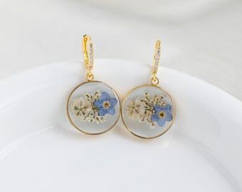 May include: A pair of gold hoop earrings with a clear round pendant featuring pressed blue and white flowers. The hoops are accented with small clear stones.
