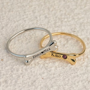 May include: Two personalised dog bone rings. One ring is silver with the inscription "Emma Max" and a heart symbol. The other ring is gold with the inscription "Kiko" and a purple gemstone.