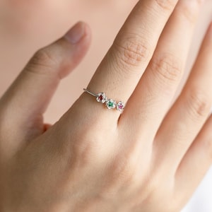 Puede incluir: Un anillo de plata con tres pequeños engastes en forma de flor, cada uno con una piedra preciosa de color diferente: rojo, verde y rosa.