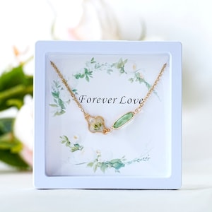 Puede incluir: Un collar de cadena dorada con un óvalo verde y un colgante de trébol de cuatro hojas que contiene una flor prensada. El collar se presenta en una caja blanca con un diseño floral y las palabras "Forever Love".