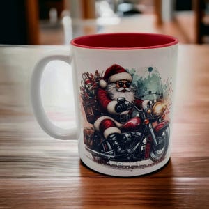 Könnte beinhalten: Eine weiße Keramiktasse mit rotem Rand. Die Tasse zeigt eine farbenfrohe Illustration von Weihnachtsmann, der ein Motorrad fährt. Das Motorrad ist rot und schwarz mit einem verchromten Scheinwerfer. Der Weihnachtsmann trägt einen roten Anzug, einen weißen Bart und eine Sonnenbrille.