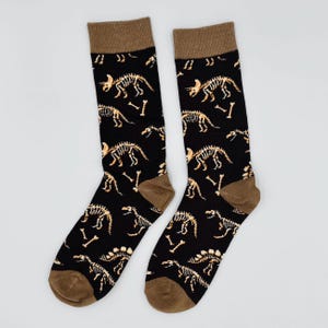 May include: A pair of black socks with a repeating pattern of dinosaur skeletons and bones in tan. The socks have brown heels, toes, and cuffs. The socks are made of a soft, comfortable material.
