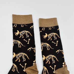 May include: Pair of black socks with a repeating pattern of dinosaur skeletons in tan and white. The socks have a tan cuff and toe. The design features various dinosaur skeletons and bone fragments.