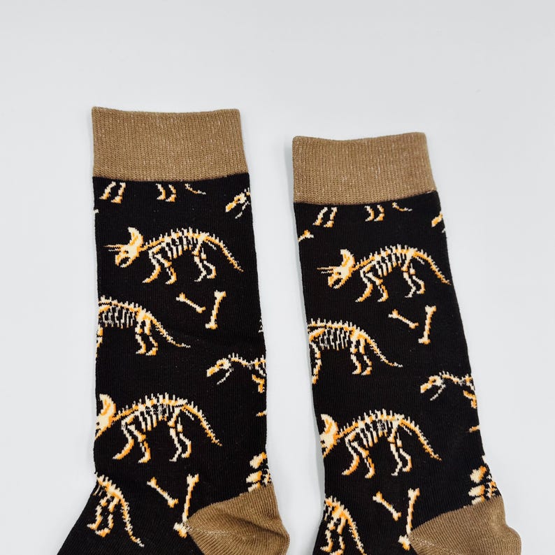 May include: Pair of black socks with a repeating pattern of dinosaur skeletons in tan and white. The socks have a tan cuff and toe. The design features various dinosaur skeletons and bone fragments.