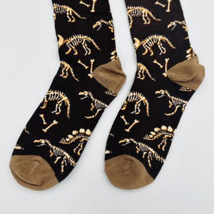 May include: A pair of black socks with a repeating pattern of dinosaur skeletons in shades of tan and brown. The socks have brown heels and toes, and are likely made of a soft, comfortable material.