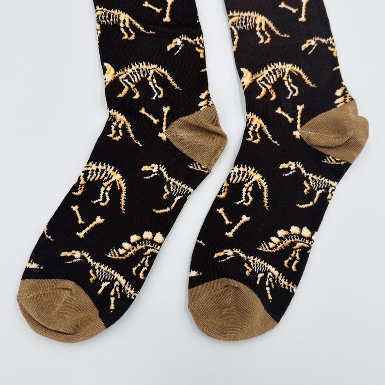 May include: A pair of black socks with a repeating pattern of dinosaur skeletons in shades of tan and brown. The socks have brown heels and toes, and are likely made of a soft, comfortable material.