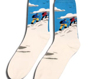 May include: A pair of crew socks featuring a blue sky with white clouds and birds above a snowy mountain landscape. The design depicts two hikers in red and blue jackets with yellow pants and backpacks climbing the mountain with trekking poles.