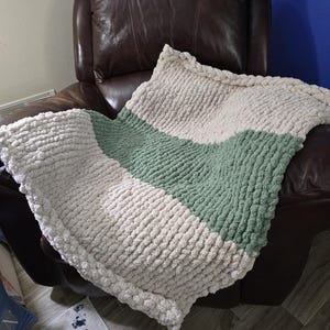 May include: A soft, chunky knit throw blanket with a cream and sage green color scheme. The blanket is draped over a brown leather armchair.