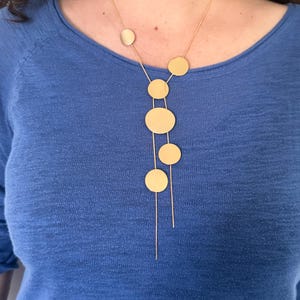 May include: Gold-toned necklace featuring a series of circular pendants of varying sizes. The necklace has a delicate chain and a modern, minimalist design. The model is wearing a blue top, creating a contrast with the gold jewelry.