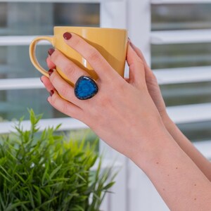 May include: A person's hand wearing a blue and black ring with a triangular shaped stone, holding a yellow ceramic mug.