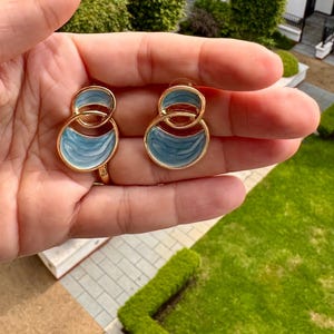 May include: A pair of gold-coloured earrings featuring a double-circle design. The upper circle is smaller than the lower one. Each circle is filled with light blue enamel with a wavy pattern. The earrings are held up against a background of green grass and a building.