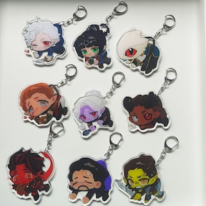 Puede incluir: Una colección de nueve llaveros acrílicos con personajes de dibujos animados en varias poses. Cada llavero tiene un anillo y una cadena de metal plateado. Los personajes tienen diferentes tonos de piel, colores de cabello y atuendos.