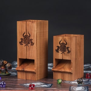 May include: Two wooden dice towers with a carved design of a horned helmet, crossed axes, and a die. The towers are different sizes and are sitting on a table with dice scattered around them.
