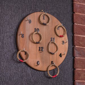 Op de afbeelding: Een houten ringwerpspel met een lichtbruine afwerking. Het spel heeft een cirkelvormig houten bord met cijfers en touwringen. De cijfers zijn zwart en de ringen zijn gemaakt van touw met rode accenten. Het spel is gemonteerd op een donkergrijze muur.