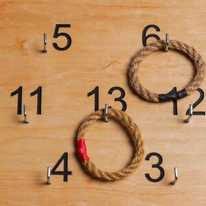 Op de afbeelding: Een houten spelbord met genummerde haken en twee touwringen. De nummers op het bord zijn 3, 4, 5, 6, 11, 12, 13. Een touwring is bruin en de andere is bruin met een rode streep.
