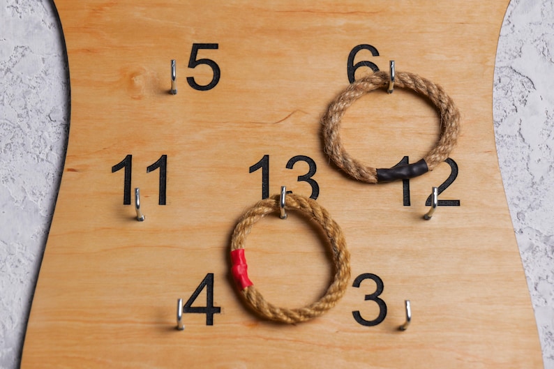 Op de afbeelding: Een houten spelbord met genummerde haken en twee touwringen. De nummers op het bord zijn 3, 4, 5, 6, 11, 12, 13. Een touwring is bruin en de andere is bruin met een rode streep.