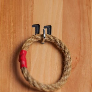 Op de afbeelding: Een houten ringwerpspel met een touwring die aan een haak hangt met het label "11". Het spel heeft ook een haak met het label "3" aan de rechterkant.