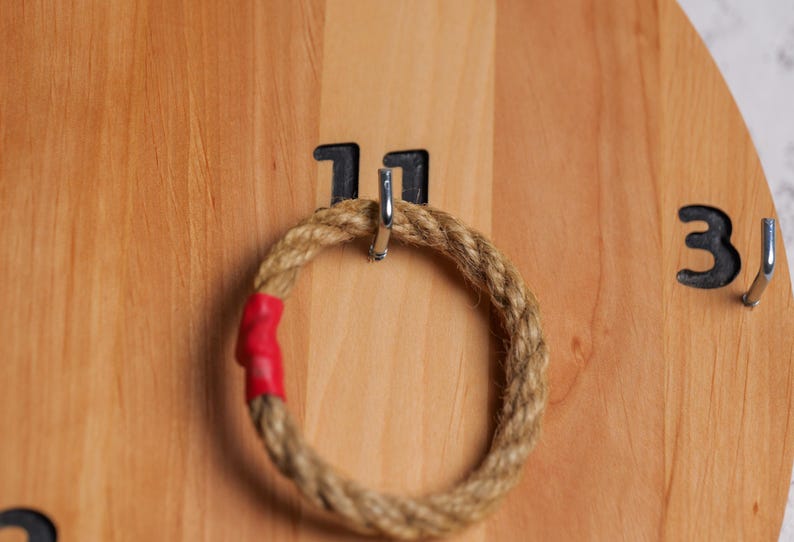 Op de afbeelding: Een houten ringwerpspel met een touwring die aan een haak hangt met het label "11". Het spel heeft ook een haak met het label "3" aan de rechterkant.