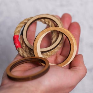 Op de afbeelding: Drie houten ringen, waarvan er &eacute;&eacute;n is omwikkeld met touw, vastgehouden in een hand. De ringen zijn van verschillende grootte en hebben een natuurlijke houten afwerking.