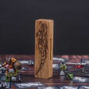 May include: A wooden dice tower with a dragon design carved into the front. The tower is made of light brown wood and has a slot on the side for rolling dice. The tower is sitting on a table with dice and other game pieces scattered around it.