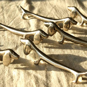 May include: A collection of metallic silver-toned drawer pulls shaped like elongated dachshund dogs. Each handle features a smooth, polished finish with distinct canine features including floppy ears, a long body, and four small legs.