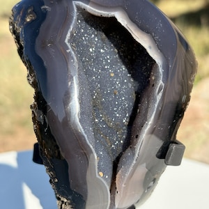 May include: A polished dark gray agate geode featuring a central cavity lined with fine, sparkling dark crystals. The exterior displays smooth, concentric banding in shades of charcoal and slate, supported by a black metal display stand.