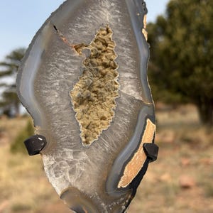 May include: A polished agate slice featuring a central geode cavity filled with small, tan-colored crystals. The stone displays concentric bands of gray, white, and translucent layers, supported by a black metal stand with two side prongs.