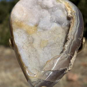 May include: A teardrop-shaped geode with a polished exterior, showcasing layers of brown, tan, and white. The interior reveals a sparkling crystal cavity. The geode is mounted on a gold-colored stand, highlighting its natural beauty.